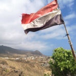 A tattered flag waving over a coastal village with mountains in the background