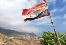 A tattered flag waving over a coastal village with mountains in the background