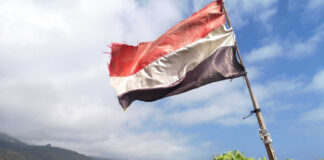 A tattered flag waving over a coastal village with mountains in the background
