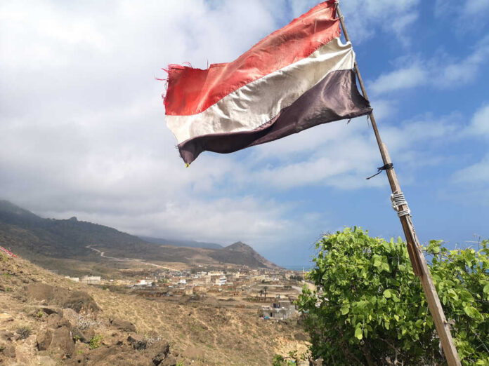 A tattered flag waving over a coastal village with mountains in the background