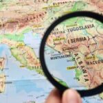 IRGC Chaos: Albania’s Move Aligns With Trump A hand holding a magnifying glass over a map highlighting Serbia and surrounding countries