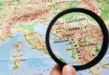 A hand holding a magnifying glass over a map highlighting Serbia and surrounding countries