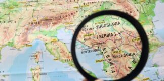 A hand holding a magnifying glass over a map highlighting Serbia and surrounding countries