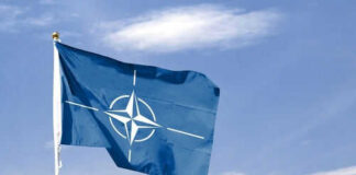 Blue NATO flag with white emblem under cloudy sky.