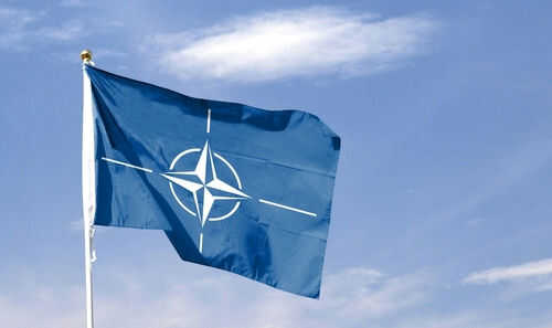 Blue NATO flag with white emblem under cloudy sky.