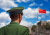 Soldier in a green military uniform facing the Great Wall of China with the Chinese flag in the background