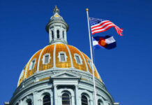 The dome of a state capitol building with flags flying against a blue sky