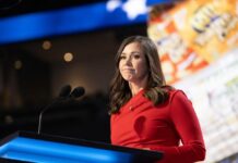 A woman in a red dress speaking at a podium with microphones