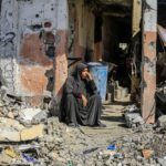 Hamas’s Sinister Scheme – Women Trapped by Aid A woman sitting among rubble outside a damaged building
