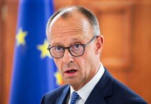 A man in a suit with glasses, looking serious in front of an EU flag