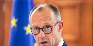 A man in a suit with glasses, looking serious in front of an EU flag
