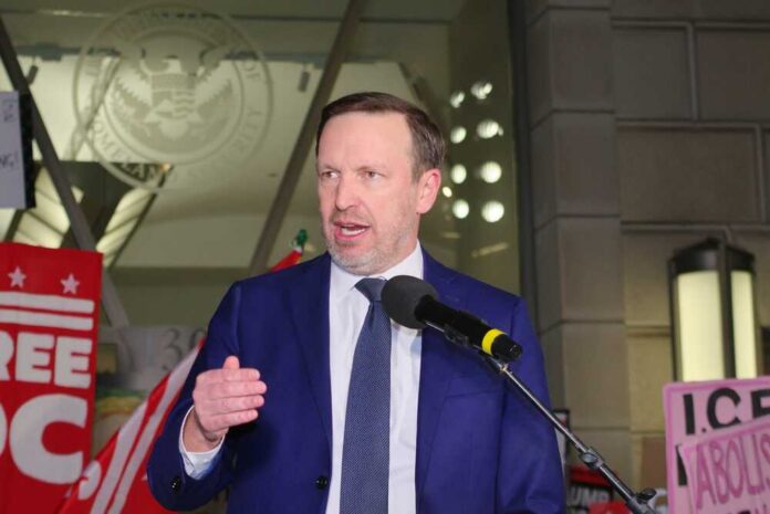 A man in a blue suit speaking at a protest with signs in the background