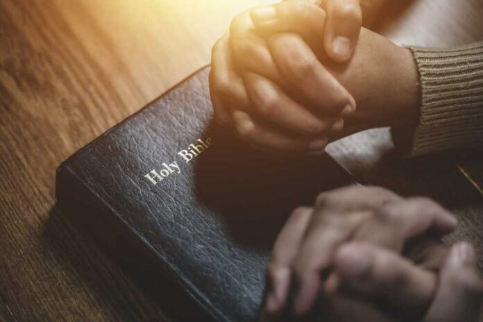 Two hands clasped in prayer over a Holy Bible