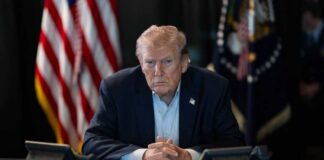 A serious-looking man seated at a table with phones and an American flag in the background