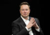 Man in black suit sitting, clasping hands.