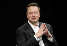 Man in black suit sitting, clasping hands.