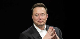 Man in black suit sitting, clasping hands.