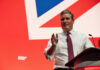 UK PM Starmer Cornered: Revolt Goes Public Man in white shirt and tie speaking at a podium on a red stage