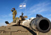 Self-Reliance Myth Meets Engine Reality Soldier saluting from atop a tank with an Israeli flag in the background