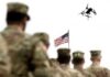 soldiers watch a drone fly near an american flag