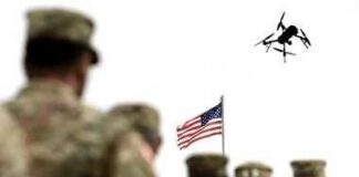 soldiers watch a drone fly near an american flag