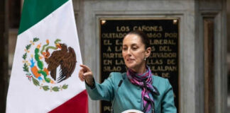 Person speaking at podium with Mexican flag nearby