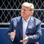 Tariff Gift? Trump’s 321-Diamond Dilemma man in a navy suit clapping in front of a chain-link fence