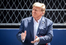 man in a navy suit clapping in front of a chain-link fence