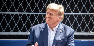 man in a navy suit clapping in front of a chain-link fence