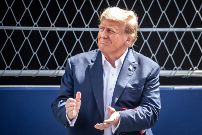 man in a navy suit clapping in front of a chain-link fence