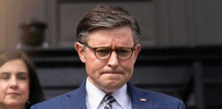 Man in blue suit with glasses looks down thoughtfully
