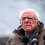 elderly man in glasses speaking outdoors into microphones