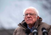elderly man in glasses speaking outdoors into microphones