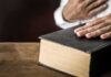 Hand on a thick black hardcover book on a wooden table