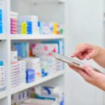 Trump Price Gambit Upends Dingell Narrative Pharmacist using a tablet in front of stocked pharmacy shelves