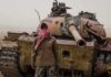 Man in scarf stands before a rusted tank in a barren field