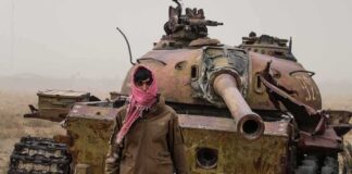 Man in scarf stands before a rusted tank in a barren field