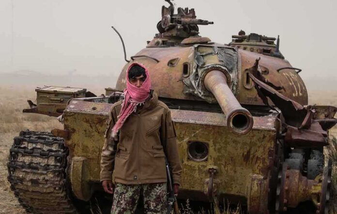 shutterstock_2027334521 Man in scarf stands before a rusted tank in a barren field