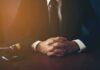 AI Fake Citations Wreck Deutsche Bank In Court person in suit with hands clasped at a desk beside a gavel