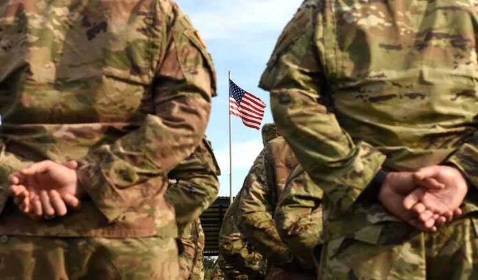 Soldiers in camouflage stand with hands behind backs facing a U.S. flag