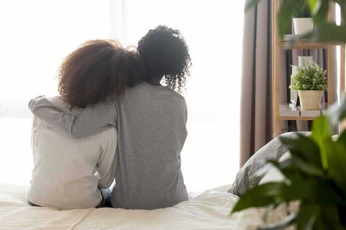 Two people sitting on a bed with arms around each other