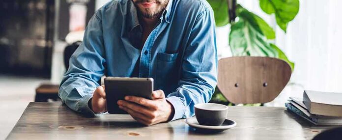shutterstock_1675165702 Man in blue shirt using a tablet at a cafe table