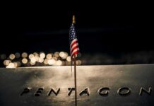 Anniversary Jolt: Trump’s Words Hit Hard Small American flag on a memorial with bokeh lights at night