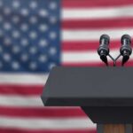 Murkowski Fumes – DOJ Crashes Alaska Count Podium with microphones in front of a blurred American flag