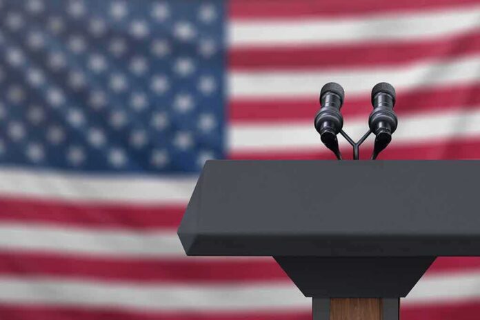 Podium with microphones in front of a blurred American flag