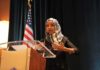 Raid Fallout or PR Stunt? Omar’s Lake Walk Ilhan Omar speaking at a podium with a microphone