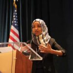 Raid Fallout or PR Stunt? Omar’s Lake Walk Ilhan Omar speaking at a podium with a microphone