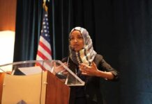 Raid Fallout or PR Stunt? Omar’s Lake Walk Ilhan Omar speaking at a podium with a microphone