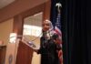 Identity Shock – Alias Bombshell Shadows Rep. Omar Ilhan Omar speaking at a podium with a microphone