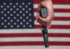 ICE Agent Allegedly Attacks – Homeowner Fires Back Hand holding a handgun in front of an American flag